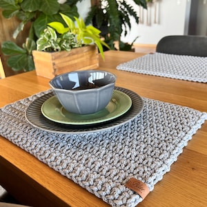 Pode incluir: Um jogo americano cinza de crochê com um padrão texturizado, combinado com uma tigela cinza, um prato verde e um prato cinza. O jogo americano está em uma mesa de madeira. Um segundo jogo americano é visível no fundo.