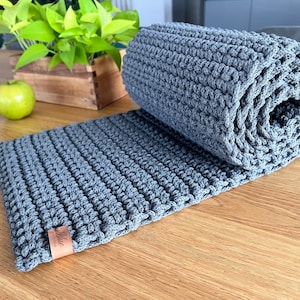 Può includere: Un runner da tavolo in crochet grigio arrotolato su un tavolo di legno. Il runner ha un'etichetta in pelle marrone con la parola "Blue" stampata su di essa.