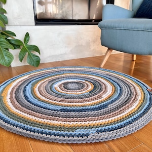 Peut inclure: Un tapis rond en crochet multicolore avec un motif de cercles concentriques dans des tons de bleu, marron, beige et blanc. Le tapis est fait de fil épais et a une surface texturée.