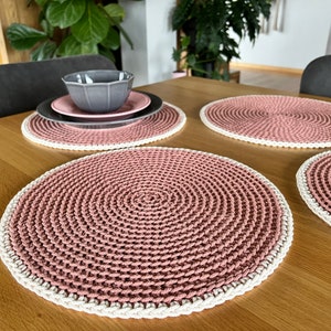 Könnte beinhalten: Vier gehäkelte Platzsets in einem staubigen Rosaton mit weißem Rand. Die Platzsets liegen auf einem Holztisch.