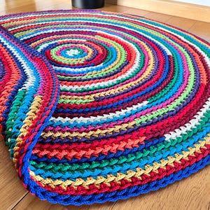 Op de afbeelding: Een rond, kleurrijk gehaakt vloerkleed met een veelkleurig spiraalpatroon. Het vloerkleed is gemaakt van dik garen en heeft een gefranst randje.