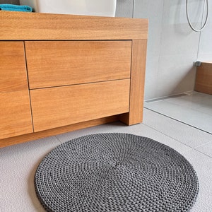 May include: A gray round crocheted bathroom rug with a brown leather tag. The rug is on a light gray tiled floor.