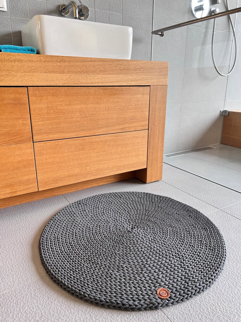 May include: A gray round crocheted bathroom rug with a brown leather tag. The rug is on a light gray tiled floor.