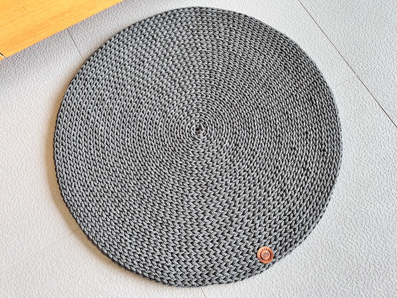 May include: A round, gray, crocheted rug with a brown leather tag in the lower right corner.