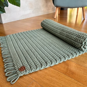 Può includere: Un tappeto verde all'uncinetto con frange sui bordi. Il tappeto è arrotolato e ha un bottone marrone sul lato.