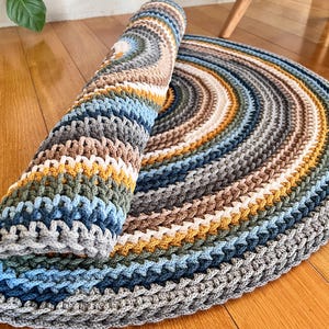 Könnte beinhalten: Ein runder, gehäkelter Teppich mit einem mehrfarbigen Streifenmuster in Grautönen, Blau, Braun und Gelb. Der Teppich ist auf der linken Seite des Bildes aufgerollt.