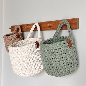 Op de afbeelding: Drie handgemaakte gehaakte manden in beige, gebroken wit en saliegroen hangen aan een houten rek. Elke mand heeft een stevige handgreep en een klein bruin leren label. De manden zijn ontworpen voor opslag.