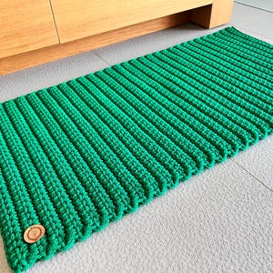 Op de afbeelding: Een groen gehaakt vloerkleed met een bruin label. Het vloerkleed heeft een gestreept patroon en is gemaakt van zacht garen.