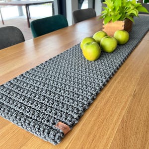 May include: A gray crocheted table runner with a textured pattern, placed on a wooden table. The runner is adorned with green apples and a small wooden box with green plants. A leather tag is attached to the runner.