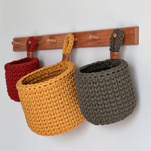 Puede incluir: Tres cestas de ganchillo hechas a mano en rojo, amarillo y gris cuelgan de un perchero de madera. Cada cesta tiene un asa de bucle con una etiqueta de cuero. Las cestas están hechas de hilo grueso y tienen una apariencia texturizada.