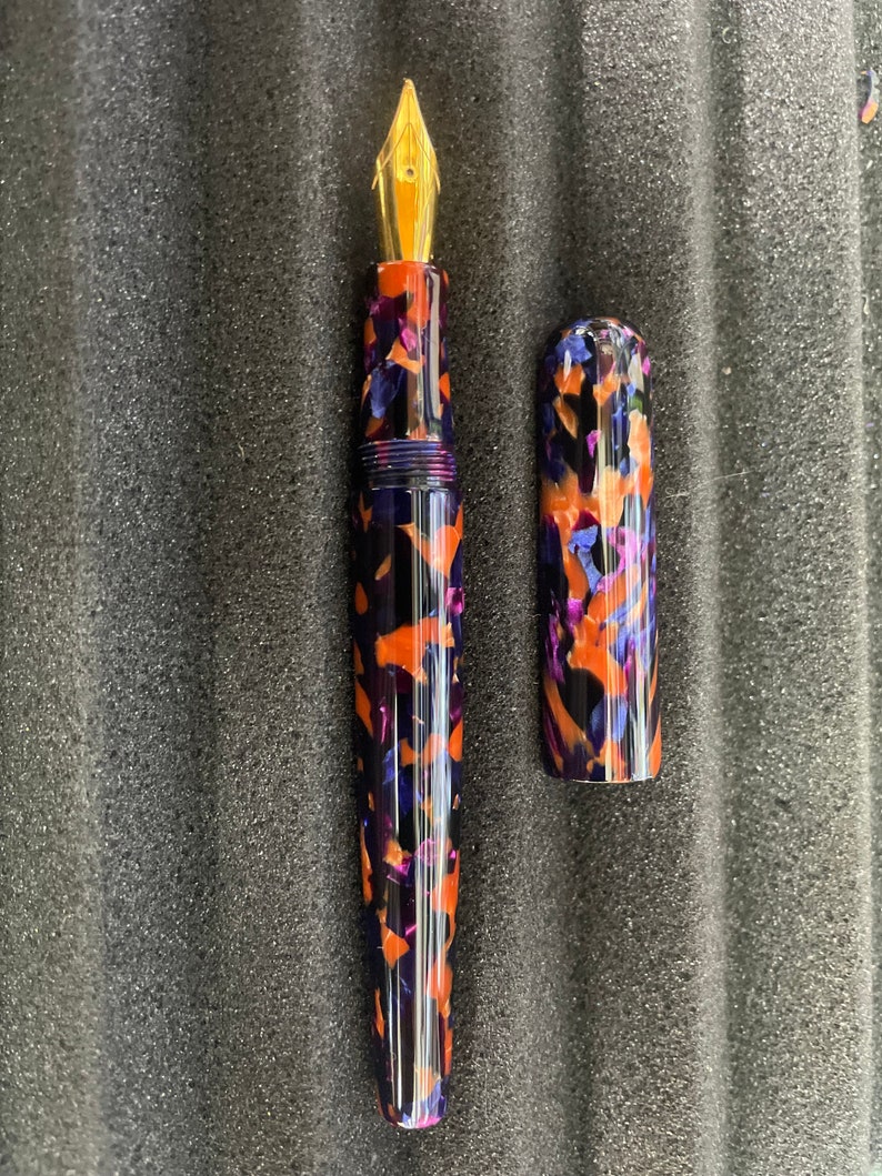 Puede incluir: Una pluma estilogr&aacute;fica negra y naranja con una punta de oro y un capuch&oacute;n a juego. La pluma tiene un patr&oacute;n de m&aacute;rmol.