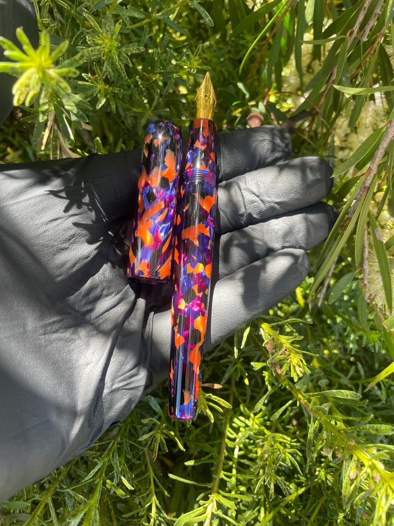 Puede incluir: Una pluma estilogr&aacute;fica negra y morada con una plumilla dorada y un patr&oacute;n abstracto colorido. La pluma est&aacute; hecha de resina y tiene una tapa que coincide con el cuerpo.