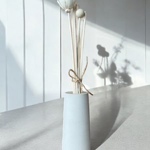 Könnte beinhalten: Eine hellblaue, geriffelte Keramikvase enthält ein Bündel dekorativer Schilf-Diffusorstäbchen. Die Stäbchen sind mit einer Blume und einer Kugel versehen und mit Bindfaden zusammengebunden. Die Vase steht auf einer hellen Oberfläche vor einer weißen Wand.