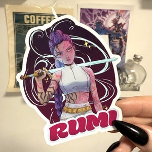 Può includere: Un adesivo con un'illustrazione stilizzata di una donna con i capelli viola che tiene una spada. L'adesivo ha un bordo bianco e la parola "RUMI" in lettere rosa. La donna ha segni rosa su braccia e viso.