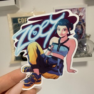 Può includere: Un adesivo con un'illustrazione stilizzata di una persona con un top blu e pantaloni gialli. L'adesivo ha il nome "Zoey" in blu con un cuore. La persona indossa scarpe da ginnastica blu e gialle.