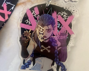 May include: A clear acrylic keychain featuring a character illustration with pink and black accents. The character has blue and gray hair, and is wearing a black top. The keychain includes a silver keyring and a black strap with pink text.