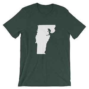 May include: Dark green t-shirt featuring a white outline of the state of Vermont with a skier in silhouette. The skier is mid-air, performing a jump. The shirt is made of a soft material.