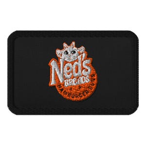 Op de afbeelding: Geborduurde patch met de tekst "Ned's Breads & Hamburgers" in oranje en wit. Het ontwerp toont een cartoon koeienkop boven de tekst, tegen een zwarte rechthoekige achtergrond.