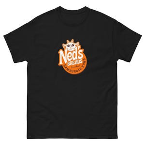 Puede incluir: Camiseta negra con un logotipo de personaje de dibujos animados para Ned's Breads. El logotipo es naranja y blanco y dice "Ned's Breads Enriched Hamburger Buns".