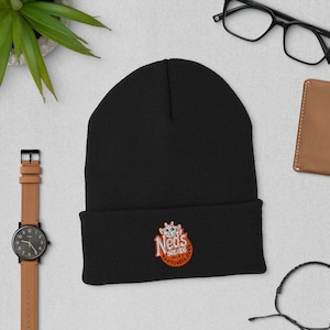 Op de afbeelding: Een zwarte beanie met een omgeslagen rand. De beanie heeft een geborduurd logo met de tekst "Ned's Breads Hamburgers" in oranje en wit. De beanie is gemaakt van een zacht, gebreid materiaal.