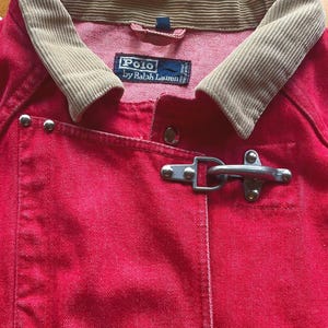 May include: A bright red denim jacket featuring a tan corduroy collar and two silver-toned metal toggle closures on the front. The interior label reads Polo by Ralph Lauren. The jacket includes silver metal rivets and a structured fabric design.