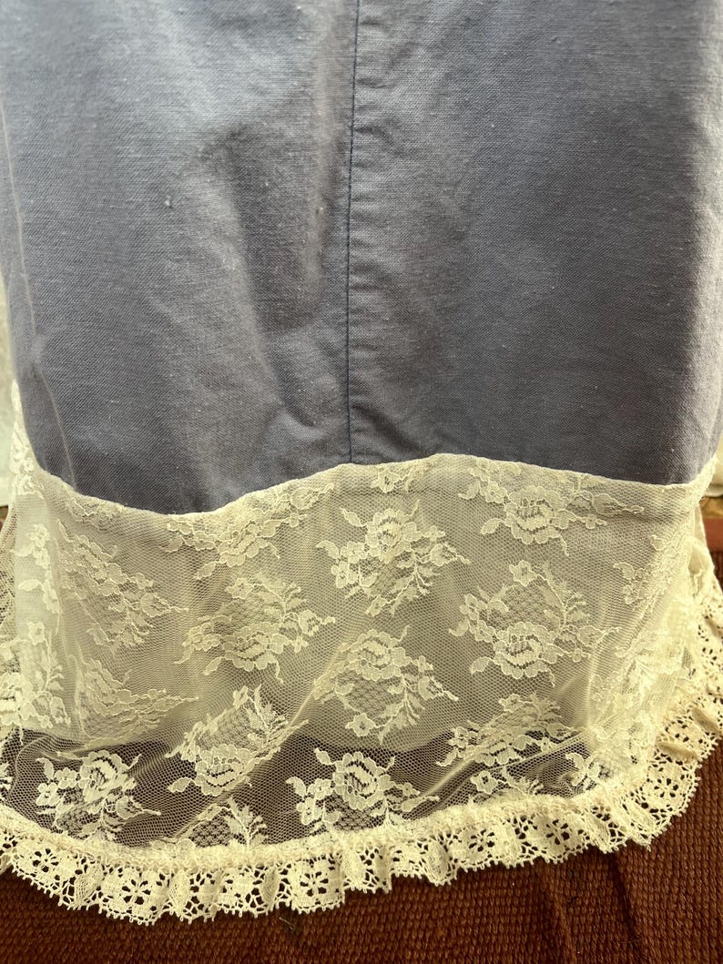 May include: A close-up of a blue fabric with a white lace trim. The lace features a floral pattern and a scalloped edge. The fabric appears to be a dress or a garment.