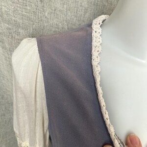 May include: Close-up of a vintage-style garment featuring a muted blue bodice and white sleeves. The bodice is trimmed with delicate cream-colored lace, and the sleeves have a sheer lace detail at the cuffs. The fabric appears soft and slightly textured.