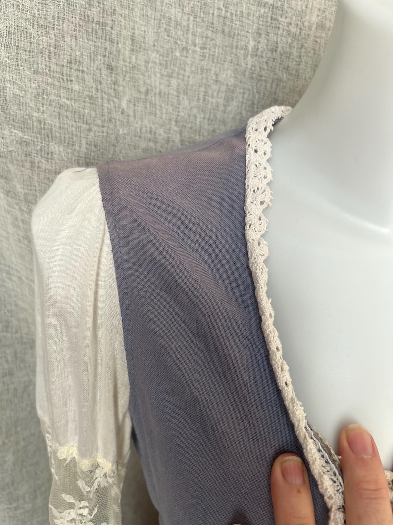 May include: Close-up of a vintage-style garment featuring a muted blue bodice and white sleeves. The bodice is trimmed with delicate cream-colored lace, and the sleeves have a sheer lace detail at the cuffs. The fabric appears soft and slightly textured.