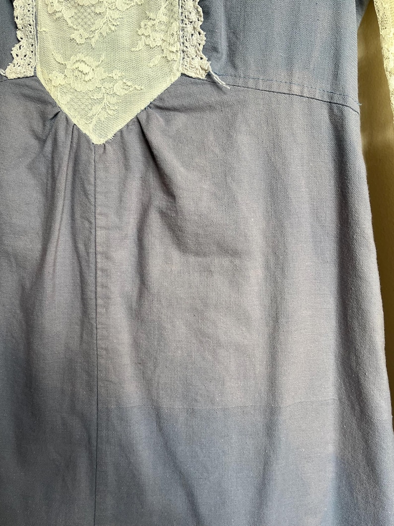 May include: Close-up of a blue dress with a white lace detail at the neckline. The lace is intricately designed with floral patterns. The dress has a simple, classic design.