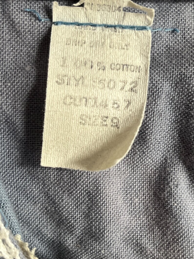May include: Close-up of a fabric tag with text. The tag is cream-colored and attached to a blue fabric. The tag reads "100% COTTON", "STY 5072", "CUT 1457", and "SIZE 9".