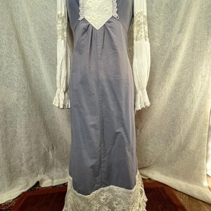 May include: A vintage-style, floor-length dress in a muted blue-gray color. The dress features white lace detailing on the bodice, sleeves, and hem. The long sleeves are white with lace accents at the cuffs. The dress is displayed on a mannequin.