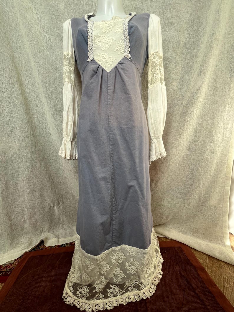 May include: A vintage-style, floor-length dress in a muted blue-gray color. The dress features white lace detailing on the bodice, sleeves, and hem. The long sleeves are white with lace accents at the cuffs. The dress is displayed on a mannequin.