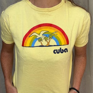 May include: A pale yellow short-sleeve t-shirt featuring a graphic print of a rainbow arching over two palm trees and a seagull. Below the rainbow, the word "cuba" is printed in a bold, dark blue lowercase font. The shirt has a crew neckline.