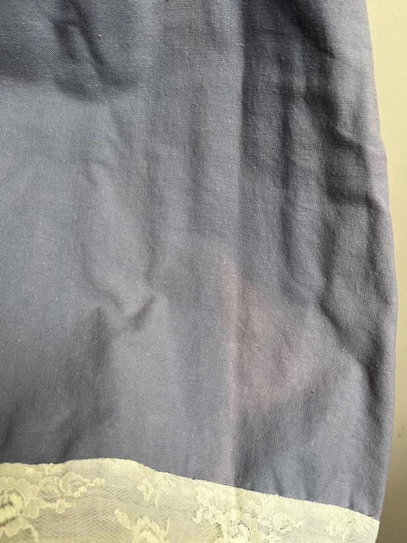 May include: Close-up of a section of fabric. The main part is a muted blue-gray color, with a lighter, off-white lace trim at the bottom. The fabric has a slightly textured appearance.