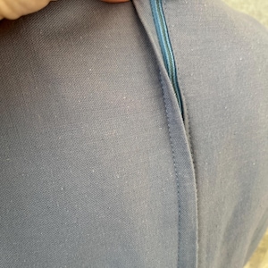 May include: Close-up of a light blue dress with a visible zipper. The zipper is a lighter shade of blue and is partially open. The dress has a textured fabric and a cream-colored lace trim.