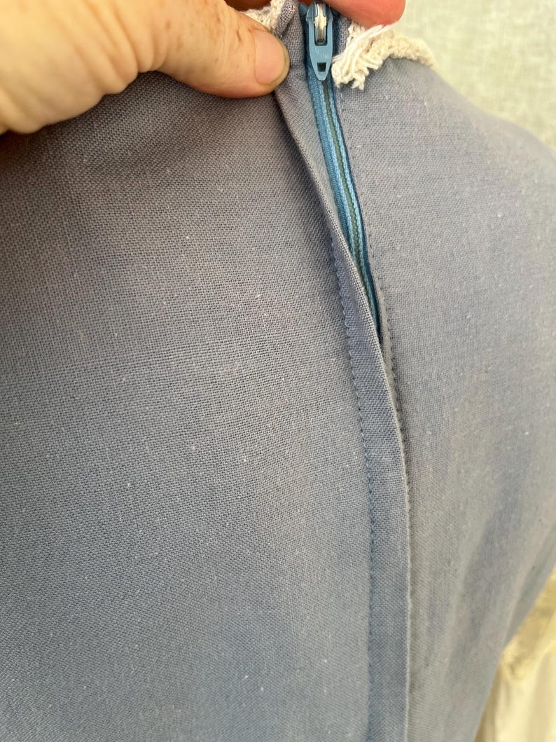 May include: Close-up of a light blue dress with a visible zipper. The zipper is a lighter shade of blue and is partially open. The dress has a textured fabric and a cream-colored lace trim.