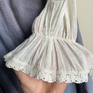May include: Close-up of a white, ruffled sleeve with a delicate crochet lace trim. The sleeve is made of a lightweight, semi-sheer fabric. The background is a soft blue fabric.