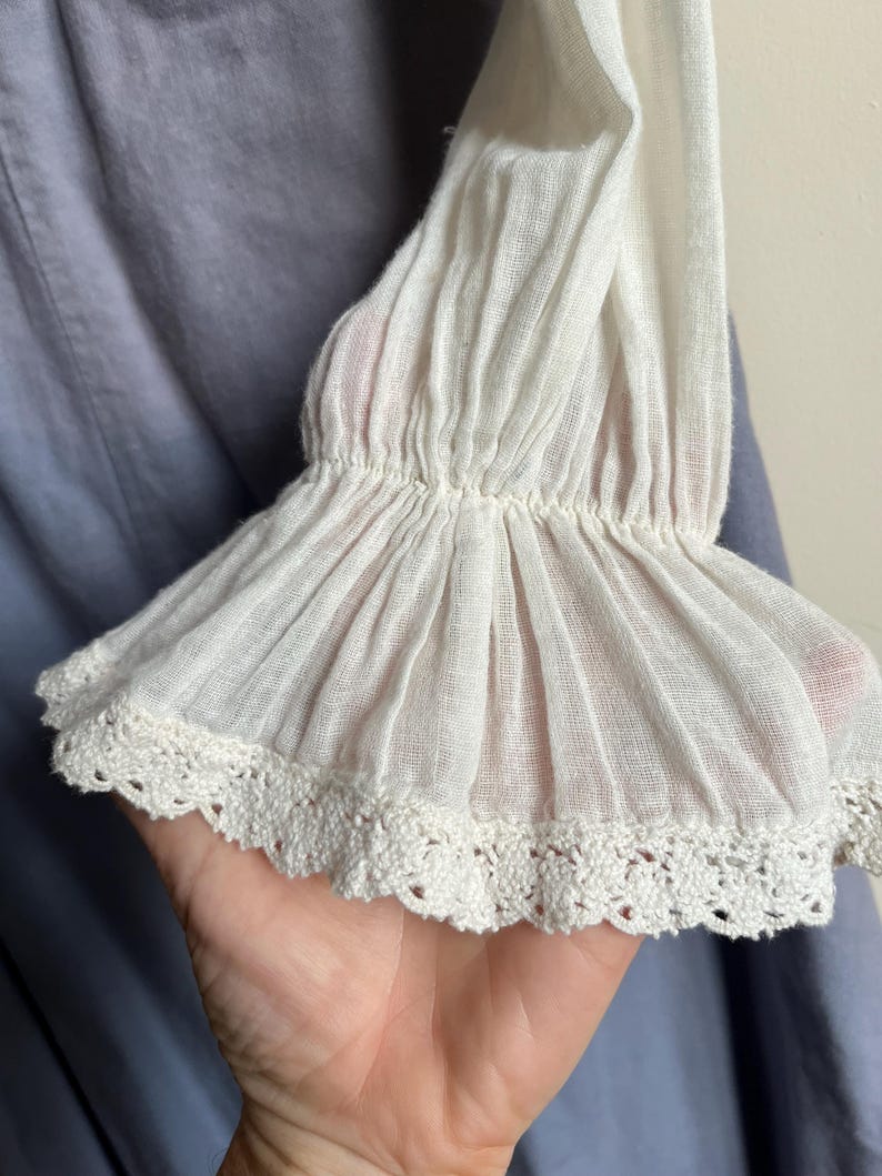 May include: Close-up of a white, ruffled sleeve with a delicate crochet lace trim. The sleeve is made of a lightweight, semi-sheer fabric. The background is a soft blue fabric.