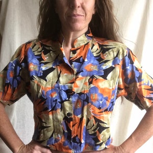 May include: A short-sleeved button-down shirt with a vibrant floral pattern. The shirt features a mandarin collar and a single pocket. The floral design includes orange, blue, and black elements on a light green background. The shirt is made of a lightweight fabric.