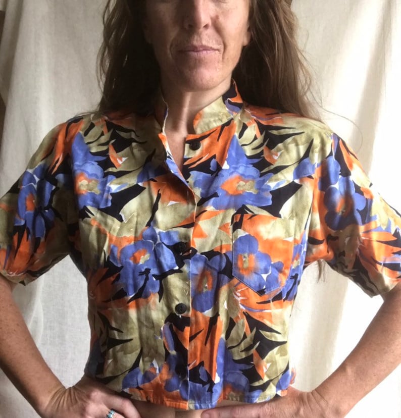 May include: A short-sleeved button-down shirt with a vibrant floral pattern. The shirt features a mandarin collar and a single pocket. The floral design includes orange, blue, and black elements on a light green background. The shirt is made of a lightweight fabric.