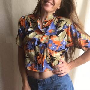 May include: A short-sleeved button-up shirt with a vibrant floral pattern in orange, blue, and black. The shirt has a mandarin collar and a single pocket. The fabric appears to be lightweight, suitable for warm weather.