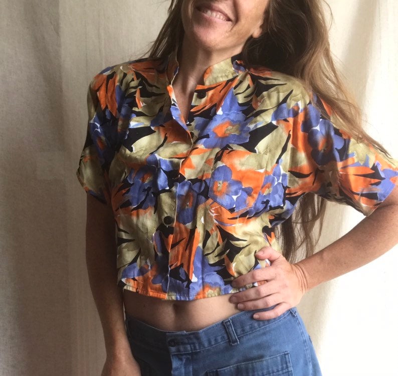 May include: A short-sleeved button-up shirt with a vibrant floral pattern in orange, blue, and black. The shirt has a mandarin collar and a single pocket. The fabric appears to be lightweight, suitable for warm weather.