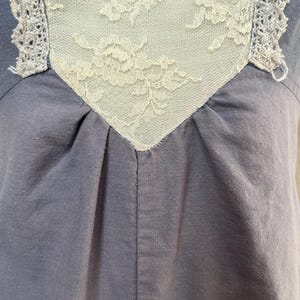 May include: Close-up of a blue-grey garment with a cream-colored lace insert. The lace is intricately patterned and framed by a delicate, crocheted trim. The fabric is gathered below the lace, creating soft folds.