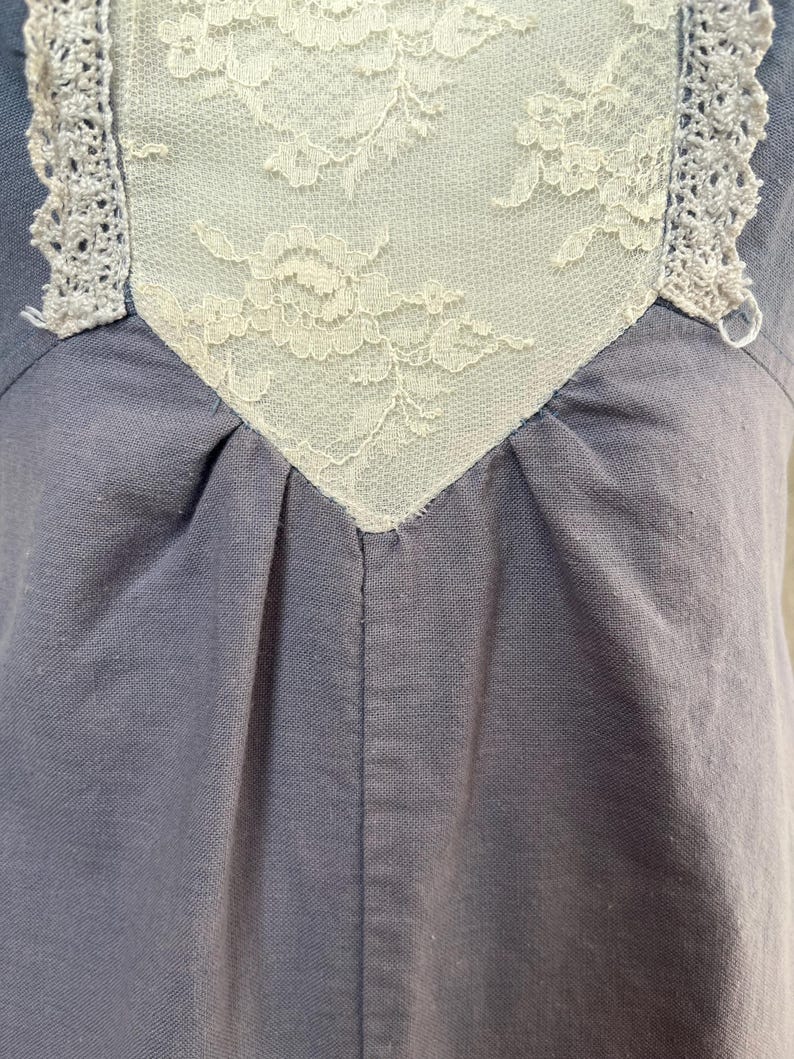 May include: Close-up of a blue-grey garment with a cream-colored lace insert. The lace is intricately patterned and framed by a delicate, crocheted trim. The fabric is gathered below the lace, creating soft folds.