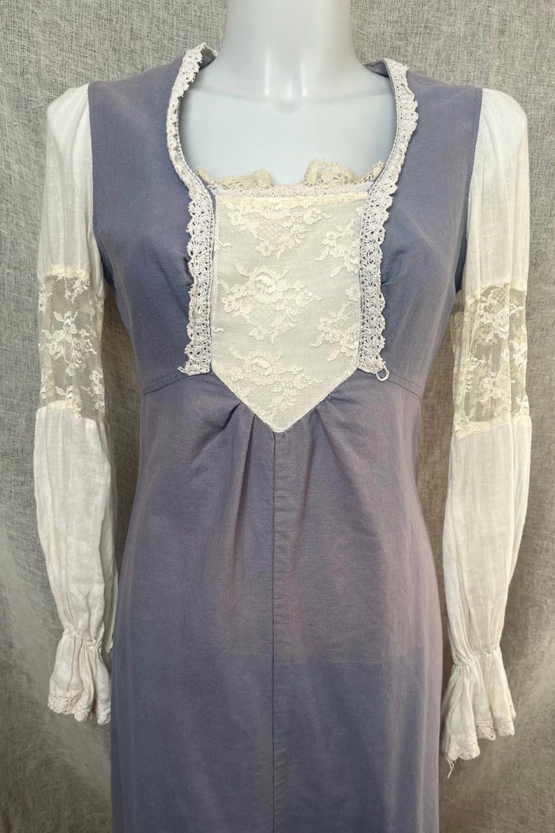May include: A vintage-style dress in a muted blue hue, featuring white lace accents. The dress has long, white sleeves with lace detailing and a square neckline trimmed with lace. The bodice is adorned with a white lace panel.
