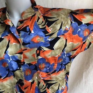 May include: Short-sleeved button-down shirt with a floral pattern. The shirt features a vibrant design with blue, orange, and black flowers on a tan background. It has a single chest pocket and a classic collar.