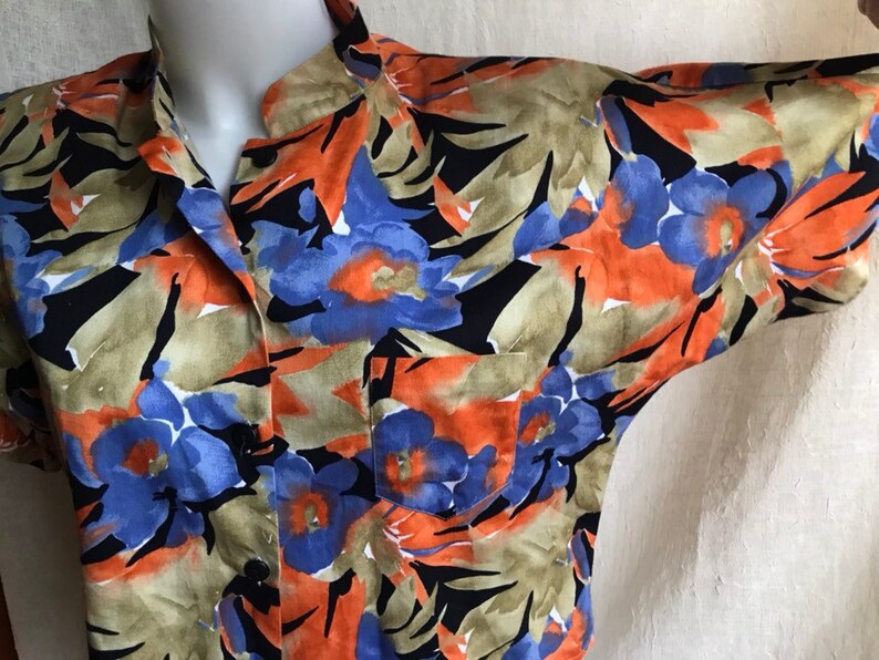 May include: Short-sleeved button-down shirt with a floral pattern. The shirt features a vibrant design with blue, orange, and black flowers on a tan background. It has a single chest pocket and a classic collar.