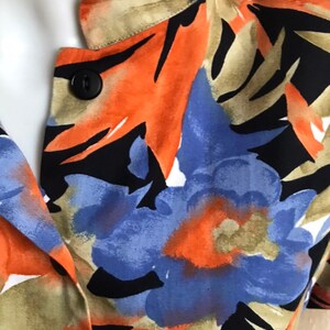 May include: Close-up of a floral print top featuring a black button. The fabric showcases a vibrant pattern of orange, blue, and black floral designs with green and tan accents. The top has a V-neckline.
