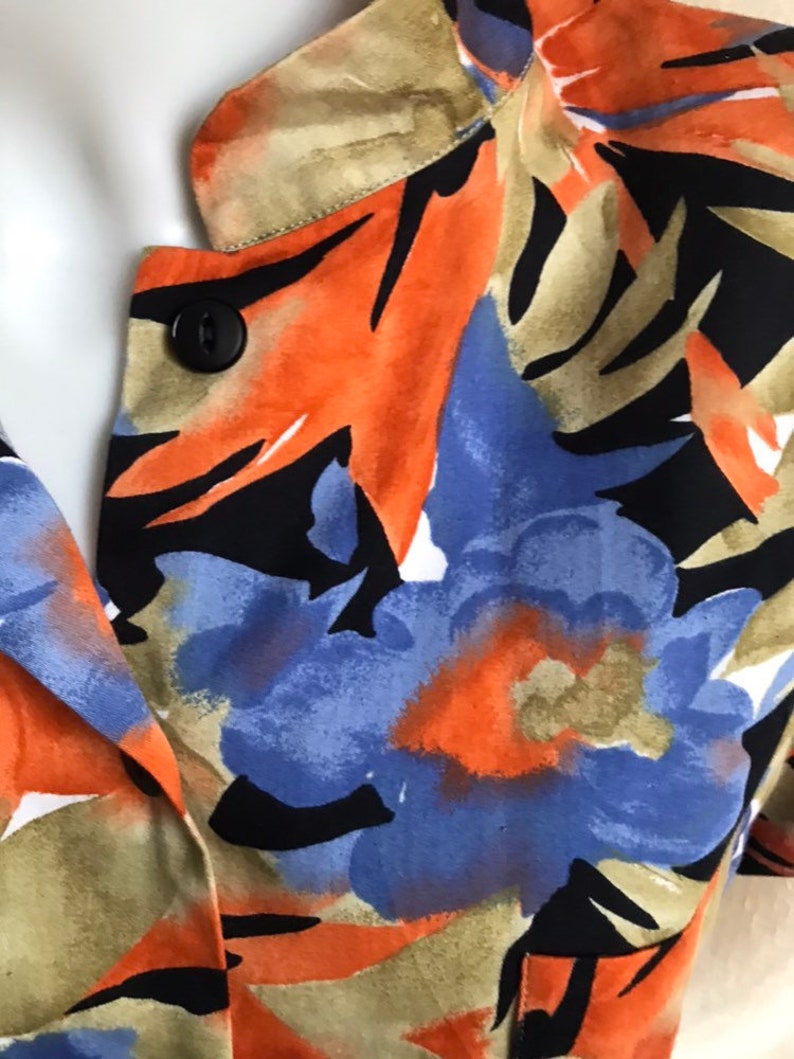 May include: Close-up of a floral print top featuring a black button. The fabric showcases a vibrant pattern of orange, blue, and black floral designs with green and tan accents. The top has a V-neckline.