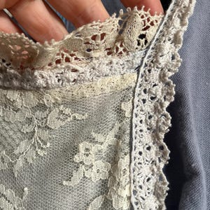 May include: Close-up of a delicate, off-white lace fabric. The lace features intricate floral patterns and is trimmed with a scalloped edge. The fabric appears to be part of a garment, possibly a vintage piece.